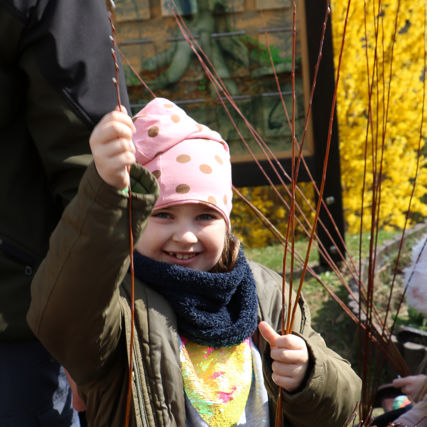 Usmívající se holčička drží v ruce proutí a bude se pokoušet plést z něj pomlázku.