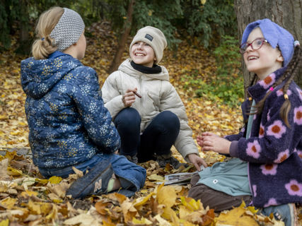 V parku Špilberk na vrstvě zlatého listí sedí tři děvčátka a s upřímným úsměvem se dívají na vedoucí kroužku (která stojí mimo záběr).