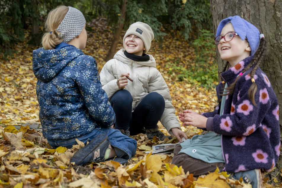 V parku Špilberk na vrstvě zlatého listí sedí tři děvčátka a s upřímným úsměvem se dívají na vedoucí kroužku (která stojí mimo záběr).
