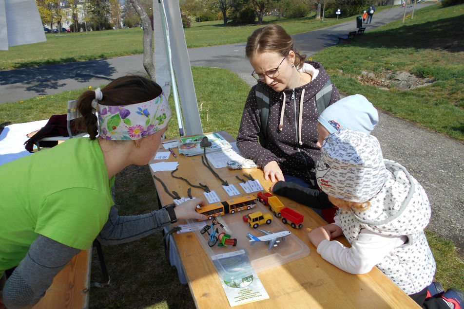 V parku na Kraví hoře sedí na lavici u stolu maminka s dcerou. Zkoumají modely různých dopravních prostředků na stole a poslouchají lektorku SEV Hlídka, která se k nim přes stůl naklání a vysvětluje uhlíkové stopy jednotlivých způsobů dopravy. 